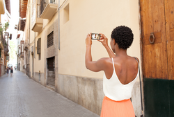 Rear view of african american tourist woman visiting a picturesque city street destination, using a smart phone taking pictures on summer holiday, outdoors. Black woman travel technology recreation.
